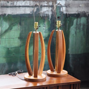 Pode incluir: Um par de candeeiros de mesa de estilo moderno de meados do s&eacute;culo com bases de madeira e detalhes em lat&atilde;o. Os candeeiros t&ecirc;m um design &uacute;nico com ripas de madeira curvas que criam um padr&atilde;o geom&eacute;trico.