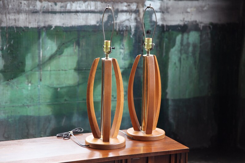 Pode incluir: Um par de candeeiros de mesa de estilo moderno de meados do s&eacute;culo com bases de madeira e detalhes em lat&atilde;o. Os candeeiros t&ecirc;m um design &uacute;nico com ripas de madeira curvas que criam um padr&atilde;o geom&eacute;trico.
