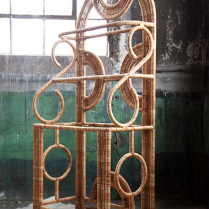 May include: A tall, free-standing, woven wicker shelf unit with a geometric design. The shelf has three tiers and is made of natural brown wicker.