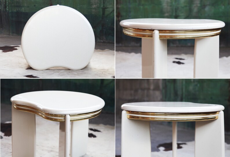 May include: White oval-shaped side table with a gold trim around the edge of the top. The table has a unique, modern design with a curved top and four legs.