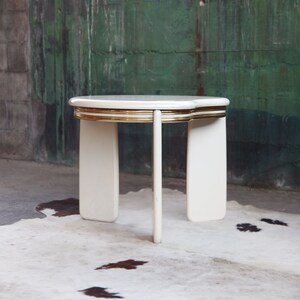 May include: White side table with a gold trim around the top edge. The table has four legs and a rounded top.