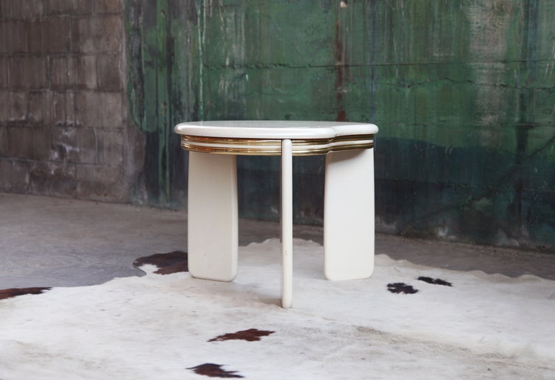 May include: White side table with a gold trim around the top edge. The table has four legs and a rounded top.