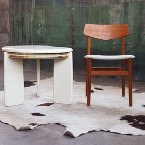May include: A white round table with a gold trim and a wooden chair with a white cushion. The table and chair are on a white and brown cowhide rug.