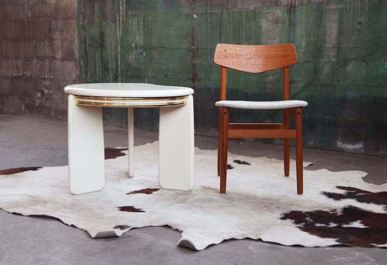 May include: A white round table with a gold trim and a wooden chair with a white cushion. The table and chair are on a white and brown cowhide rug.