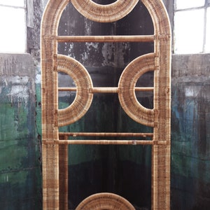 May include: A tall, arched, woven wicker shelf unit with a geometric design. The shelf has three levels and is made of natural, light brown wicker.