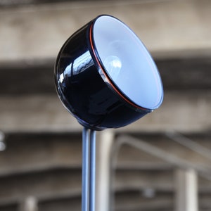 Może przedstawiać: Czarna i srebrna lampa biurkowa z okrągłym, białym kloszem. Lampa ma długi, cienki trzon i podstawę z czarnym wykończeniem.