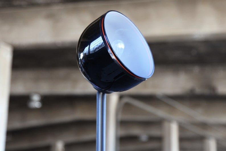 Może przedstawiać: Czarna i srebrna lampa biurkowa z okrągłym, białym kloszem. Lampa ma długi, cienki trzon i podstawę z czarnym wykończeniem.