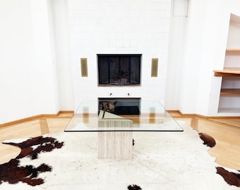 May include: A glass-top coffee table with a travertine base sits on a white cowhide rug in a living room with a fireplace. The fireplace has a white tile surround and a black metal grate.