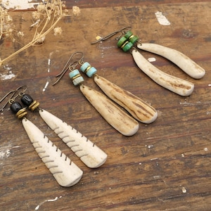 May include: Handmade earrings with a bohemian style. The earrings feature bone-shaped pendants with carved details, accented with beads in various colors. The ear wires are a dark bronze color. The earrings are displayed on a rustic wooden surface.
