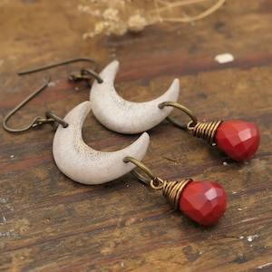 May include: A pair of silver crescent moon earrings with red gemstone drops. The earrings are made of metal and have a rustic, bohemian style.