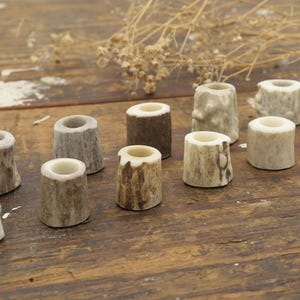 May include: A collection of small, cylindrical beads crafted from natural deer antler. Each bead features a unique, rugged exterior texture with varying shades of brown, tan, and cream, and a smooth, hollow center hole for threading.