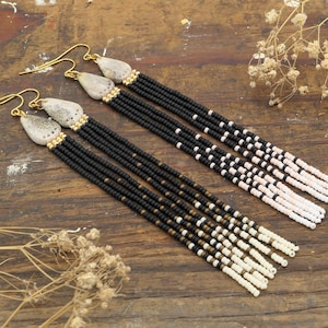 May include: A pair of long, beaded earrings with a natural shell charm. The earrings are made with black, white, and gold beads and have a tassel-like design.