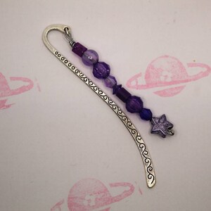 May include: A silver bookmark with a purple star charm and purple glass beads. The bookmark has a decorative swirl design.