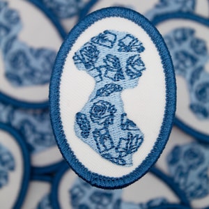 May include: A close-up of a blue and white embroidered patch featuring a floral design in the shape of a person's silhouette. The patch is oval-shaped and has a blue border.
