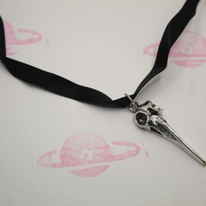 May include: A silver bird skull pendant on a black ribbon choker necklace.