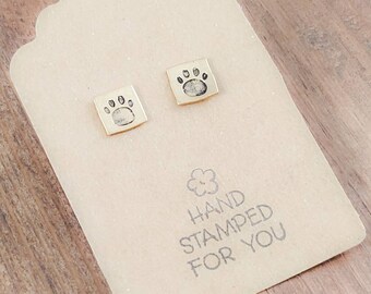 May include: A pair of gold square earrings with a paw print design. The earrings are on a brown card with the text "HAND STAMPED FOR YOU".