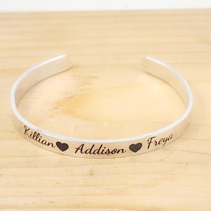 May include: A silver cuff bracelet with the names "Killian", "Addison", and "Freya" engraved on it, separated by heart symbols. The bracelet is open-ended and sits on a light wood surface.