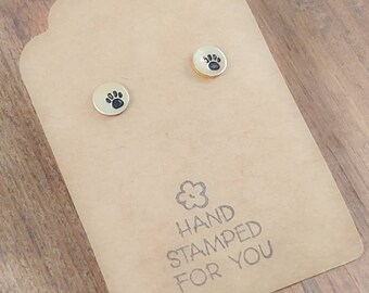 May include: A pair of gold-toned earrings with a paw print design. The earrings are displayed on a brown card with the text "HAND STAMPED FOR YOU".