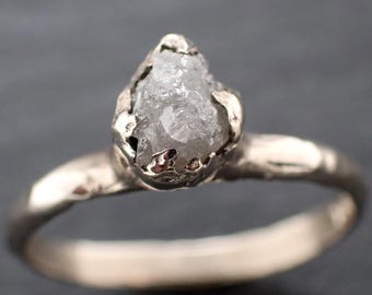 May include: A silver ring featuring a rough-cut, light gray diamond. The diamond is set in a textured, organic-looking silver band. The ring's design emphasizes the natural shape of the gemstone, creating a unique piece of jewelry.