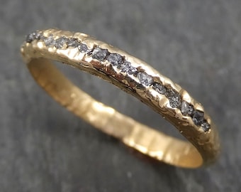 Può includere: Un anello d'oro con una fascia strutturata e una fila di piccole pietre grigio scuro. L'anello ha un aspetto martellato, che gli conferisce un aspetto rustico e unico. Le pietre sono incastonate lungo la parte superiore della fascia.