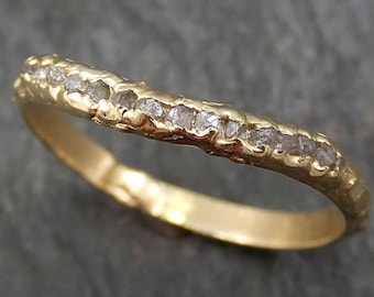 May include: A curved gold band ring featuring a series of small, rough-cut, translucent white diamonds set along the top edge. The metal surface has a textured, organic finish, and the band follows a gentle wave shape across its length.