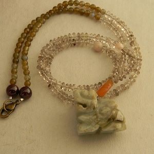May include: A necklace with a carved jade pendant, featuring a light green color. The necklace is made of multiple strands of brown, clear, and pink beads.