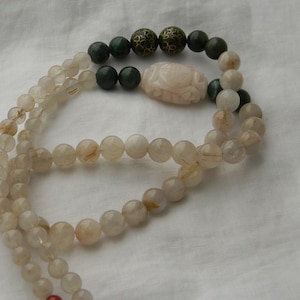 May include: A long necklace made of clear quartz beads with gold flecks. The necklace has a carved pink stone pendant and green jade beads.