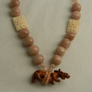 May include: A pink and white beaded necklace with a carved wooden dragon pendant. The beads are round and have a smooth, polished finish. The dragon is detailed and has a fierce expression.