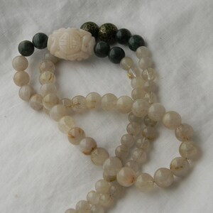 May include: A long necklace made of clear quartz beads with gold flecks. The necklace has a white jade pendant with a carved design and green jade beads at the ends.