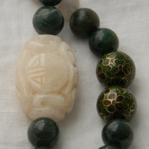 May include: A white jade bead with a carved design, surrounded by green jade and green enamel beads with gold floral patterns.