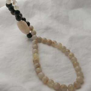May include: A long necklace made of light brown beads with gold flecks. The necklace is on a white fabric background.