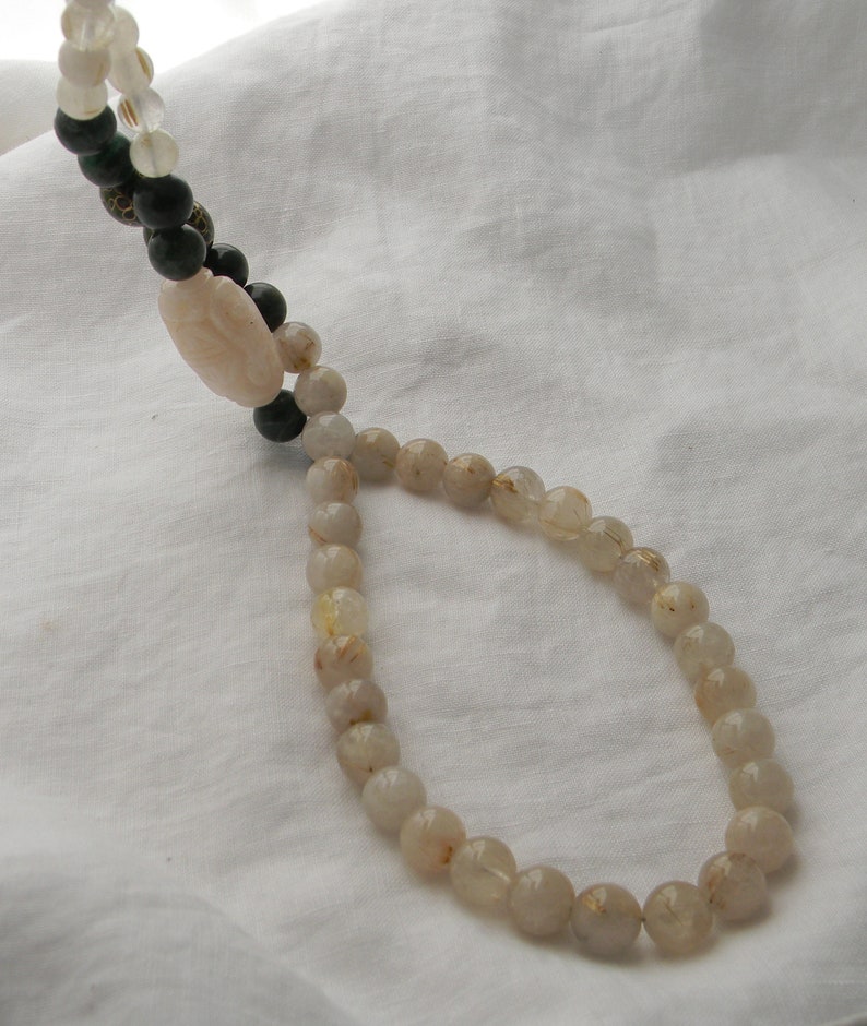 May include: A long necklace made of light brown beads with gold flecks. The necklace is on a white fabric background.