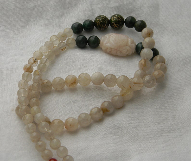 May include: A long necklace made of clear quartz beads with gold flecks. The necklace has a carved pink stone pendant and green jade beads.