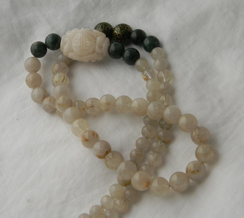May include: A long necklace made of clear quartz beads with gold flecks. The necklace has a white jade pendant with a carved design and green jade beads at the ends.