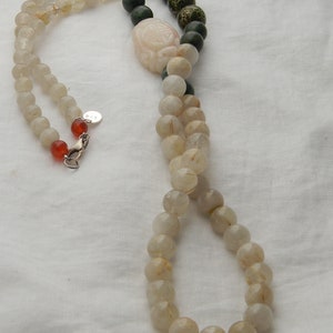 May include: A long necklace made of clear quartz beads with gold flecks, green jade beads, and a carved jade pendant. The necklace has a silver clasp.