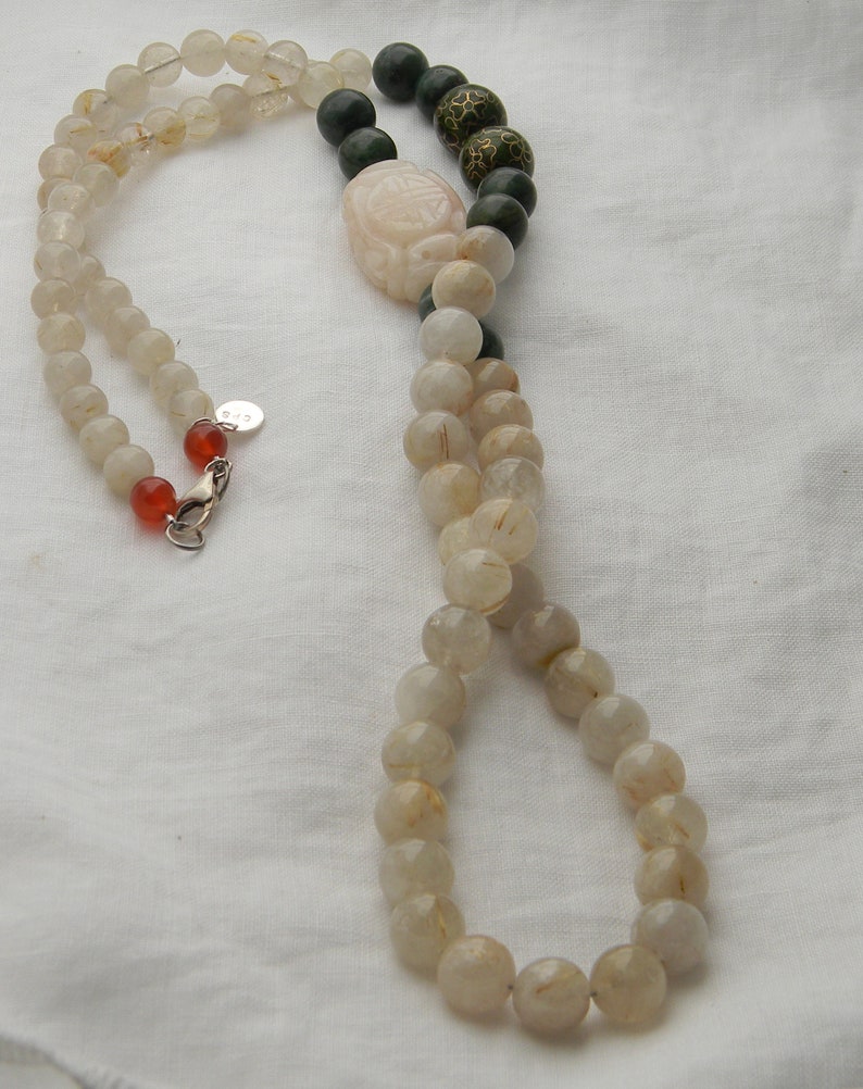 May include: A long necklace made of clear quartz beads with gold flecks, green jade beads, and a carved jade pendant. The necklace has a silver clasp.