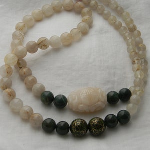 May include: A double strand necklace made of clear quartz beads with gold flecks. The necklace has a carved white jade pendant and green jade beads with gold accents.