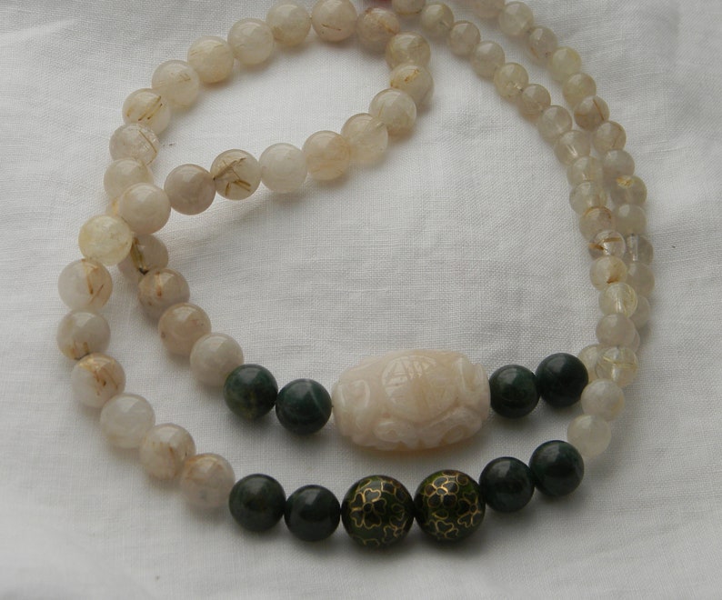 May include: A double strand necklace made of clear quartz beads with gold flecks. The necklace has a carved white jade pendant and green jade beads with gold accents.