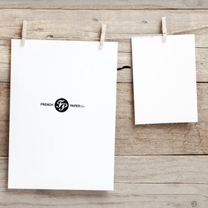 K&ouml;nnte beinhalten: Zwei wei&szlig;e Papierbl&auml;tter h&auml;ngen an einer Holzwand mit W&auml;scheklammern. Das Papier links tr&auml;gt den Text "French FP Paper Co.". Das Papier rechts ist leer.