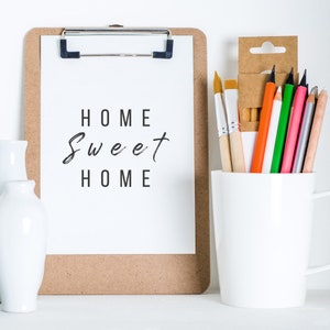 K&ouml;nnte beinhalten: Eine Klemmbrett mit Korkr&uuml;cken und einem wei&szlig;en Blatt Papier, auf dem der Text "Home Sweet Home" in Schwarz geschrieben ist. Eine wei&szlig;e Keramiktasse mit einer Auswahl an Buntstiften und Pinseln.