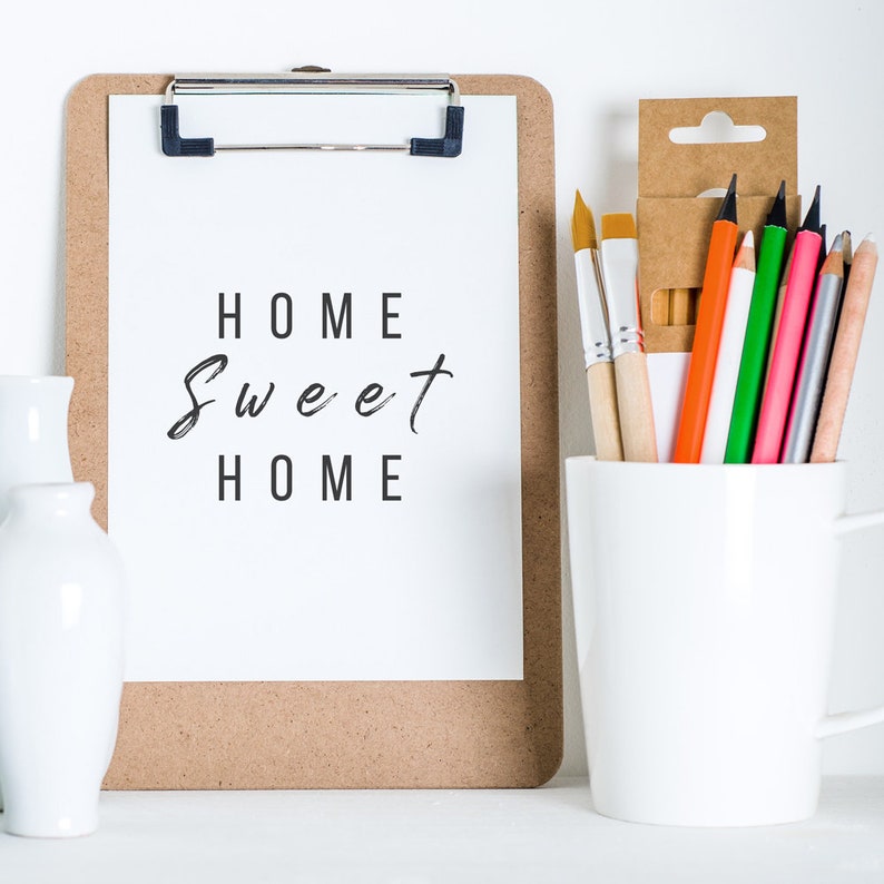 K&ouml;nnte beinhalten: Eine Klemmbrett mit Korkr&uuml;cken und einem wei&szlig;en Blatt Papier, auf dem der Text "Home Sweet Home" in Schwarz geschrieben ist. Eine wei&szlig;e Keramiktasse mit einer Auswahl an Buntstiften und Pinseln.