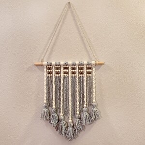 May include: A grey and white macrame wall hanging with wooden dowel and white beads. The hanging features tassels at the bottom.