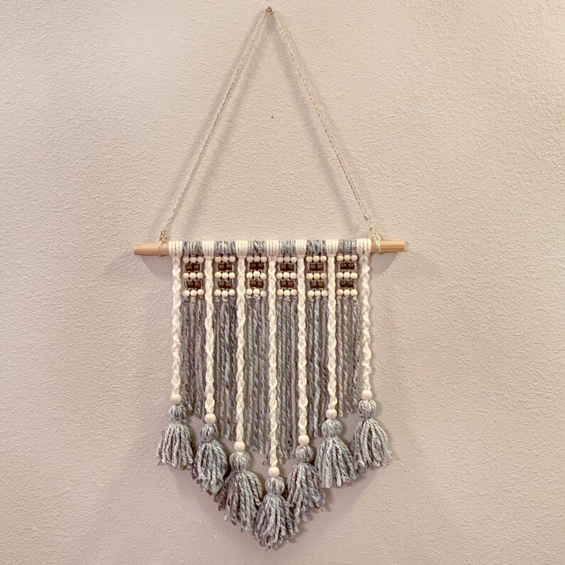 May include: A grey and white macrame wall hanging with wooden dowel and white beads. The hanging features tassels at the bottom.