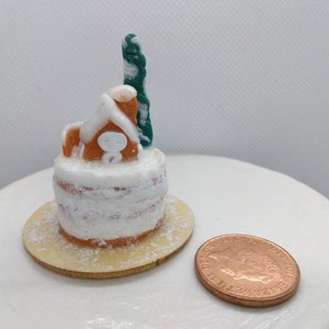 May include: A miniature gingerbread house cake with white icing and a green icing tree on top. The cake is on a small wooden base with powdered sugar sprinkled around it. A British penny is in the foreground for scale.
