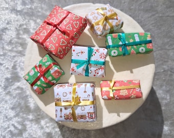 May include: A collection of seven miniature gift boxes wrapped in festive holiday paper featuring patterns of candy canes, gingerbread men, and ornaments. Each small rectangular package is tied with a thin satin ribbon in red, green, or gold.