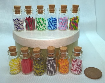 May include: Twelve small glass jars with cork tops filled with colorful candy canes. The jars are arranged in two rows of six on a white surface.