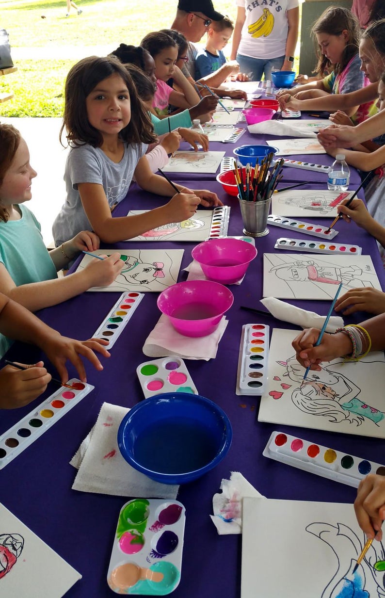 May include: A group of children are painting on canvases at a table covered in a purple tablecloth. They are using watercolors and paintbrushes. There are several bowls of water and paint palettes on the table.