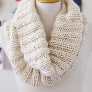 Puede incluir: Bufanda infinita de punto grueso de color crema, exhibida en un maniquí. La bufanda está hecha de hilo grueso con una textura acanalada, creando una apariencia acogedora y cálida. El color neutro la convierte en un accesorio versátil.