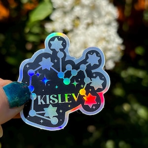 May include: A holographic sticker featuring a constellation design and the word "Kislev" in black lettering. The sticker has a rainbow iridescent finish.