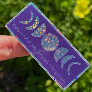 Può includere: Un adesivo rettangolare viola con un bordo olografico. L'adesivo presenta disegni di fasi lunari e il testo "Hashem loves you through every phase."